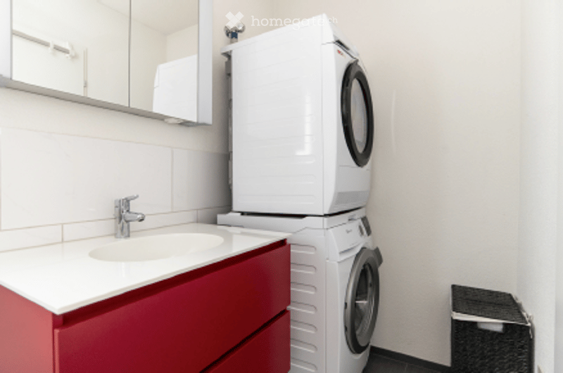 Modern bathroom with a red vanity, white sink, mirror, and stacked washing machine and dryer.