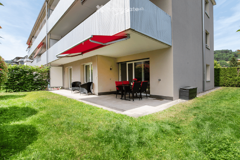Well-maintained garden with a patio area featuring a dining set, lounge chairs, and a red awning extending from the building.