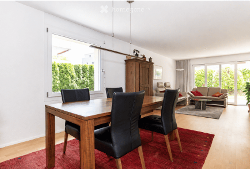 Bright, open-plan living and dining area with large windows, wooden flooring, a red area rug, a dining table with black chairs, a sofa, a coffee table, and a wooden cabinet.