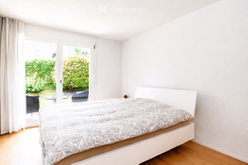 Bright bedroom with wooden flooring, white walls, large window with white curtains, bed with patterned duvet, and a chair near the window.