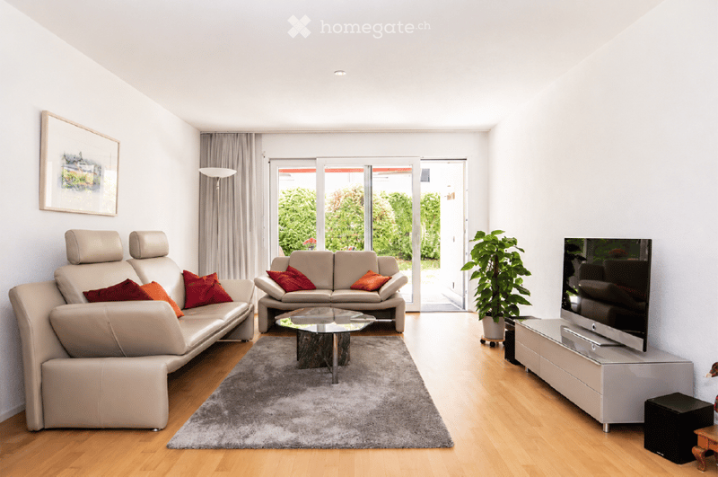Modern living room with light-colored leather sofas, a glass coffee table, a flat-screen TV, and a potted plant near the sliding glass doors that lead to a garden view.