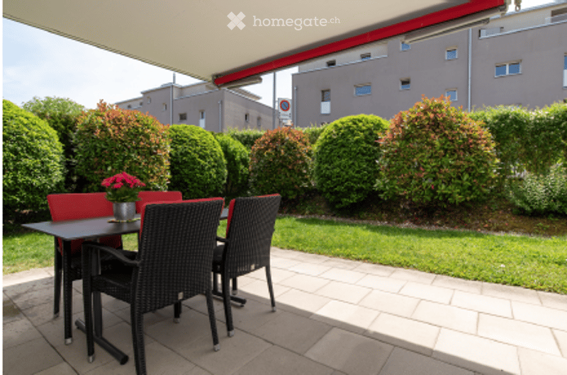 Outdoor patio with a table and chairs, surrounded by neatly trimmed bushes and a well-maintained lawn, with a building in the background.