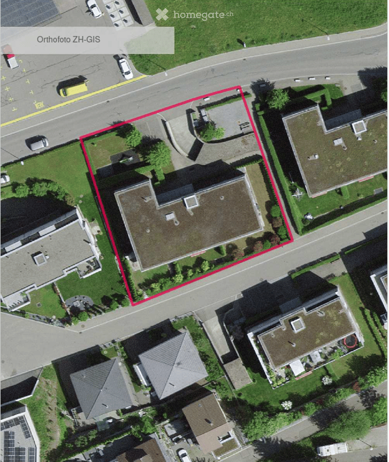 Aerial view of a residential building with a flat roof, surrounded by greenery and a parking area.