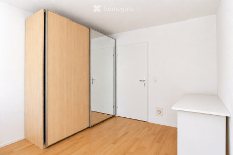 Completely empty room with white walls, wooden floor, large mirrored wardrobe, and a white cabinet.