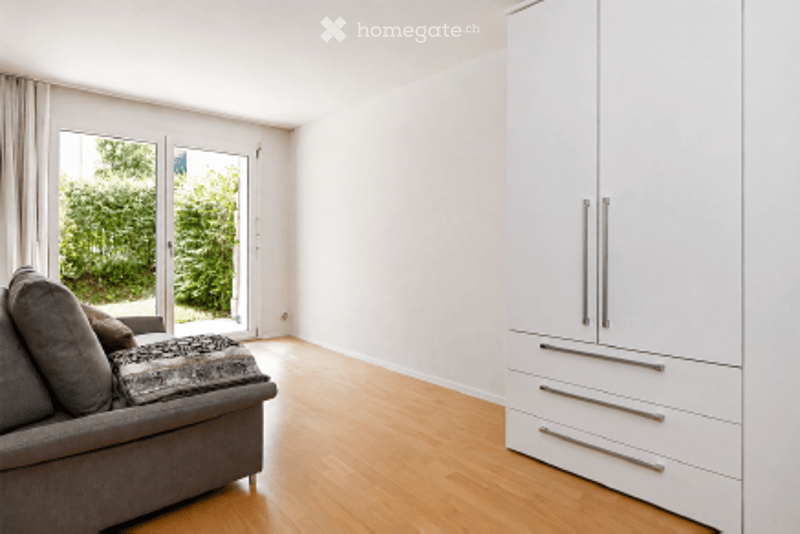 Bright living room with large glass doors leading to a garden, grey sofa, and white wardrobe with silver handles.