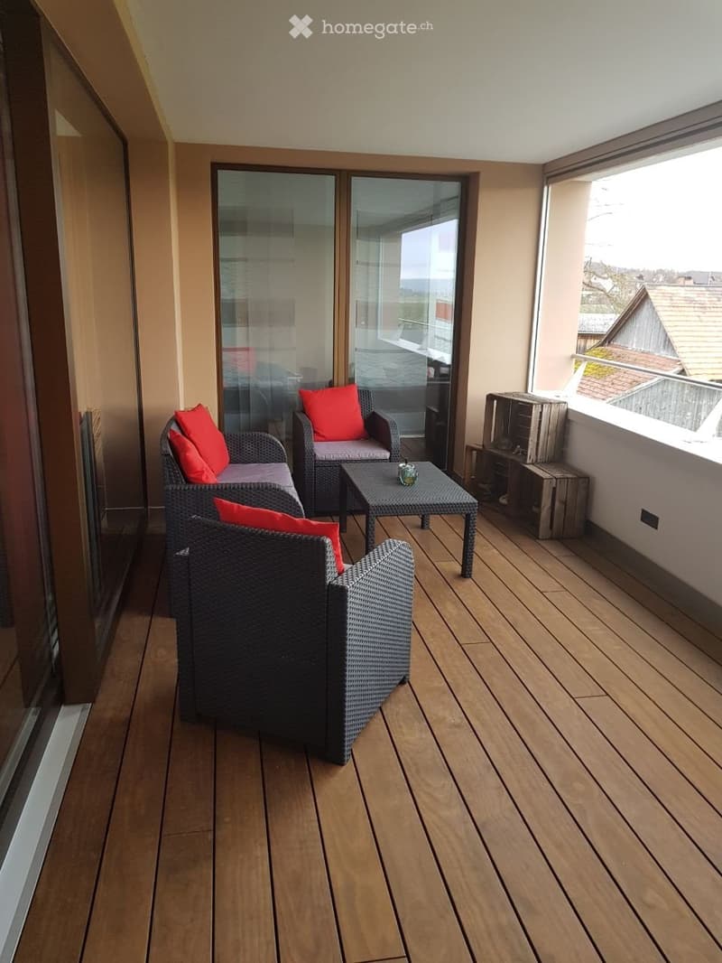 Balcony with wooden decking, wicker sofa and chairs with red cushions, small table with a cup, and a view of the surrounding area.
