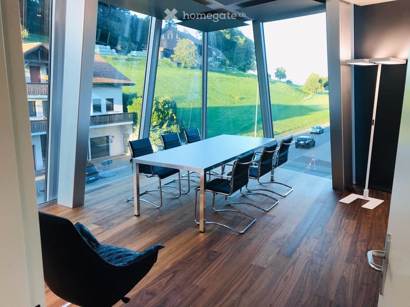 Modern office or meeting room with a white table, black chairs, and a sleek black leather chair, large windows overlooking a green landscape.