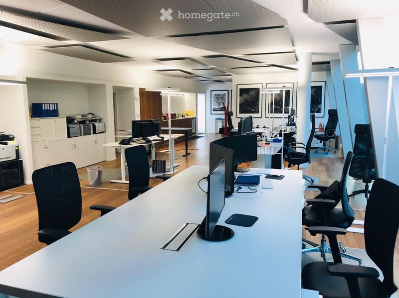 Modern office space with multiple desks, office chairs, computer monitors, keyboards, and mice. There are printers, cabinets, and framed pictures on the walls.