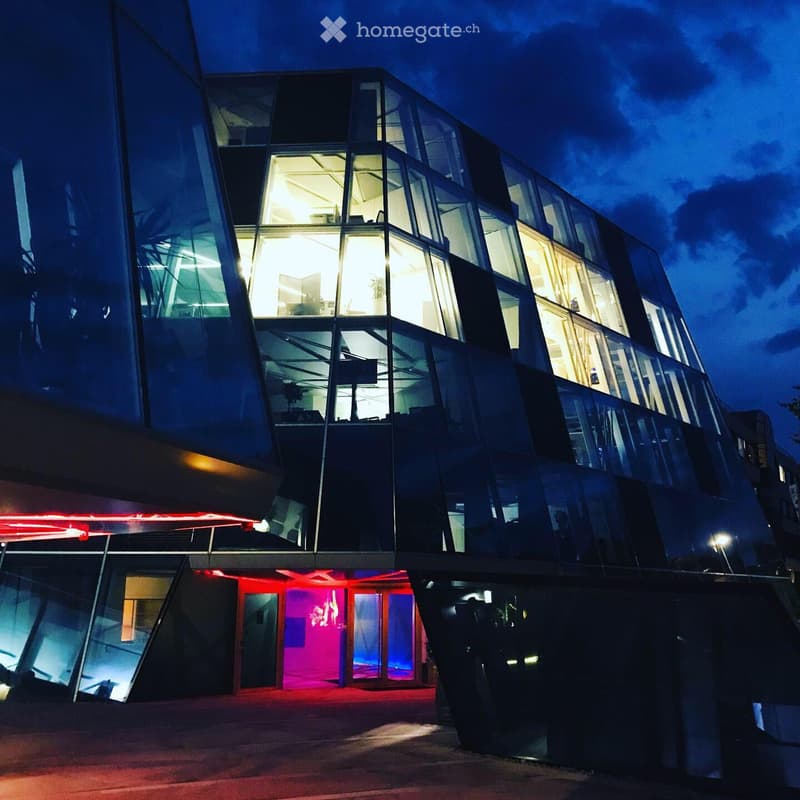 Modern, multi-story building with a curved, glass facade illuminated at night. Visible interior lighting and a vibrant red-lit entrance area.