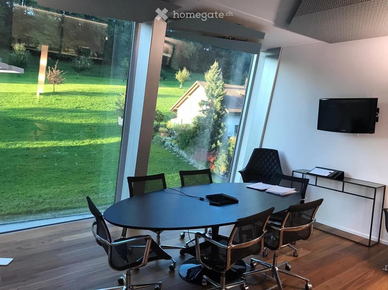 Modern office or meeting room with a round black table, several black office chairs, a wall-mounted TV screen, and large windows overlooking a green garden.