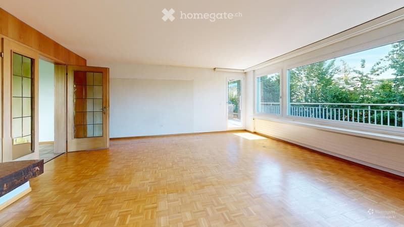 Empty room with wooden herringbone flooring, large windows with views of greenery, and a balcony accessible through a door.