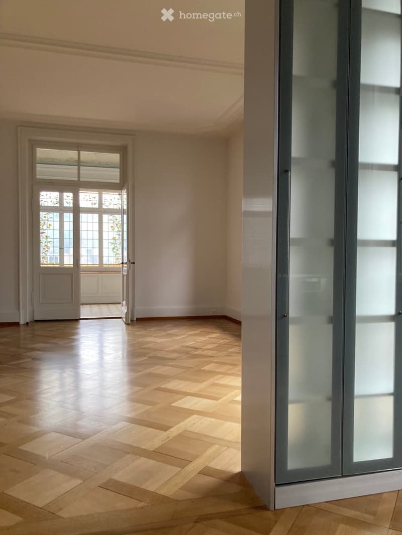 Completely empty room with wooden herringbone flooring, white walls, and large windows with frosted glass panels. No furniture or equipment.