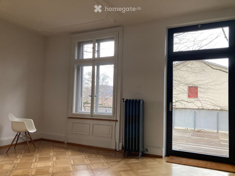 Empty room with wooden flooring, large windows, radiator, and a single white chair near the window.