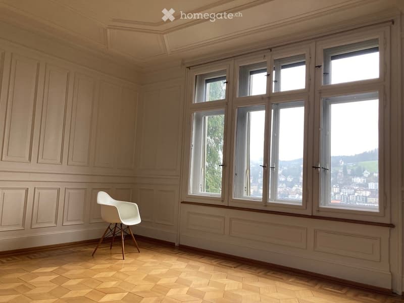 Bright, unfurnished room with large windows offering a scenic view, featuring a single white chair and wooden flooring.