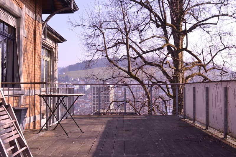 Balcony with wooden decking, metal railing, table, and chair, overlooking a tree and distant cityscape.