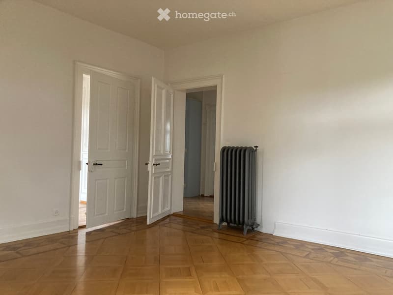 Empty room with wooden flooring, white walls, and a radiator. Two white doors are visible, one leading to another room.
