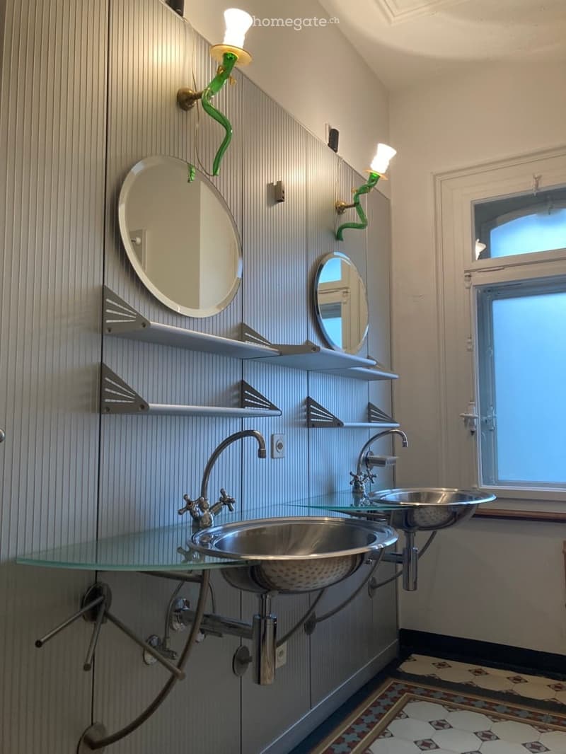 Modern bathroom with two stainless steel sinks on a glass countertop, two mirrors, decorative wall-mounted light fixtures, and white shelves.