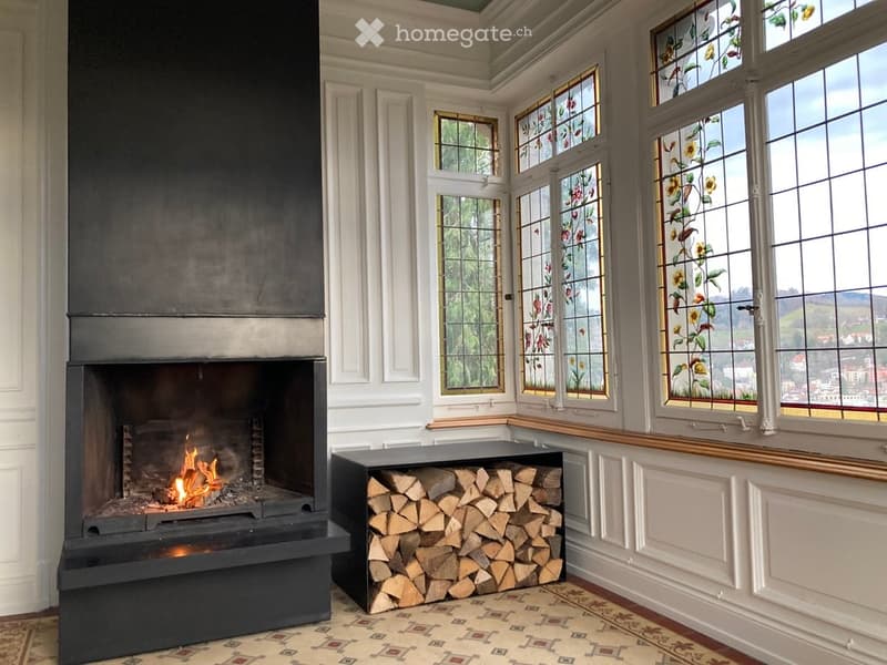 Elegant living room with a large, black fireplace, a neatly stacked wood pile, and large stained-glass windows offering a scenic view.