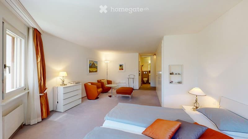 Cozy bedroom with a double bed, nightstand, lamp, and a seating area with orange chairs and a small table. The room features white walls, a large window with curtains, and a view of a hallway leading to a bathroom.