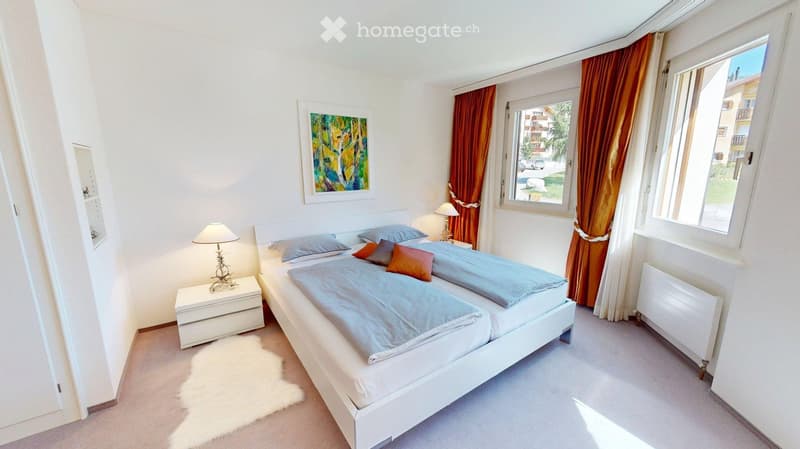 Bright bedroom with a double bed, nightstand, lamp, picture frame, and radiator. The room features large windows with orange curtains and a view of the outside.