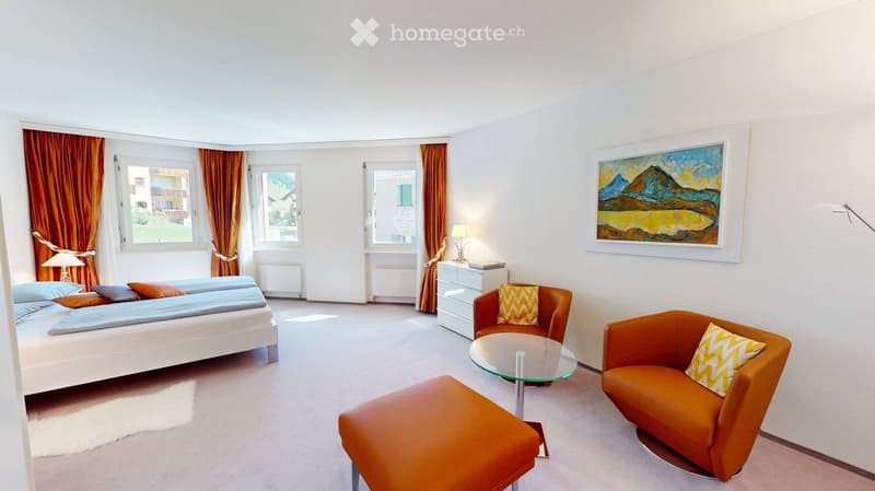 Bright bedroom with a white bed, nightstand, two orange chairs, a glass coffee table, and a painting on the wall. The room has large windows with orange curtains and a view of the outside.