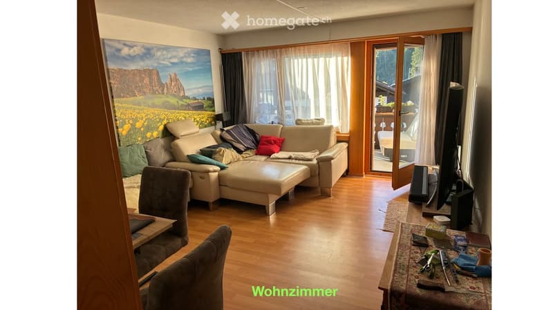 Bright living room with wooden flooring, large windows, and a balcony access. Features a beige sectional sofa with multiple cushions, an ottoman, a grey armchair, a small table, and a television set.