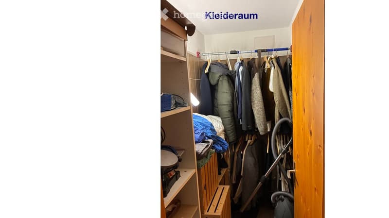 A well-organized closet with wooden shelves, a hanging rod with various garments, and a vacuum cleaner placed on the floor.