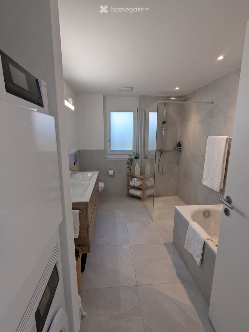 Modern bathroom with a bathtub, double sink, toilet, and walk-in shower. Features include a towel rack, wooden shelf with rolled towels, and a large window with frosted glass.