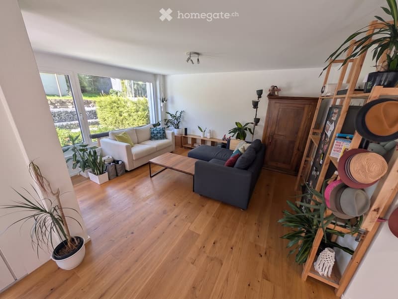 Bright living room with light wooden flooring, white walls, large windows, two sofas, coffee table, side table, floor lamp, and several potted plants.