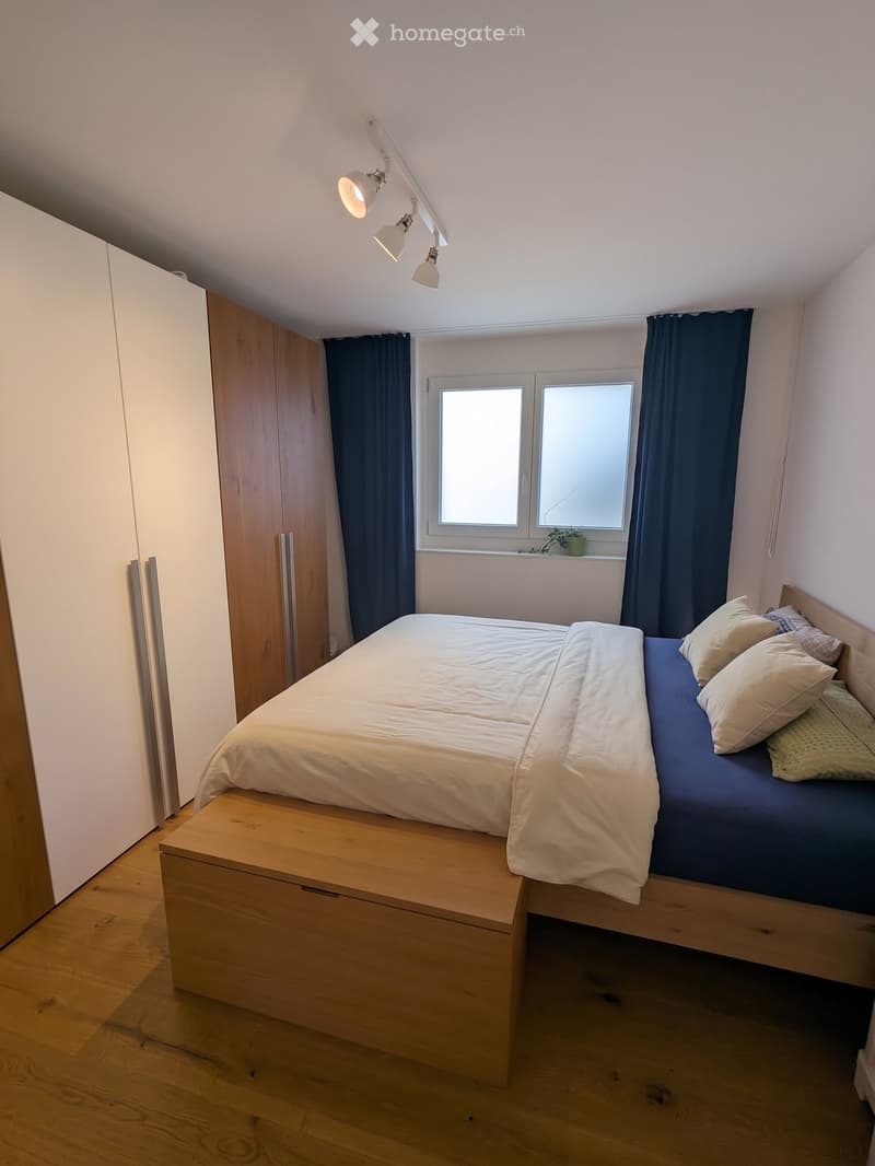 A modern bedroom with a wooden bed, white bedding, multiple pillows, a wardrobe, and a window with dark curtains.