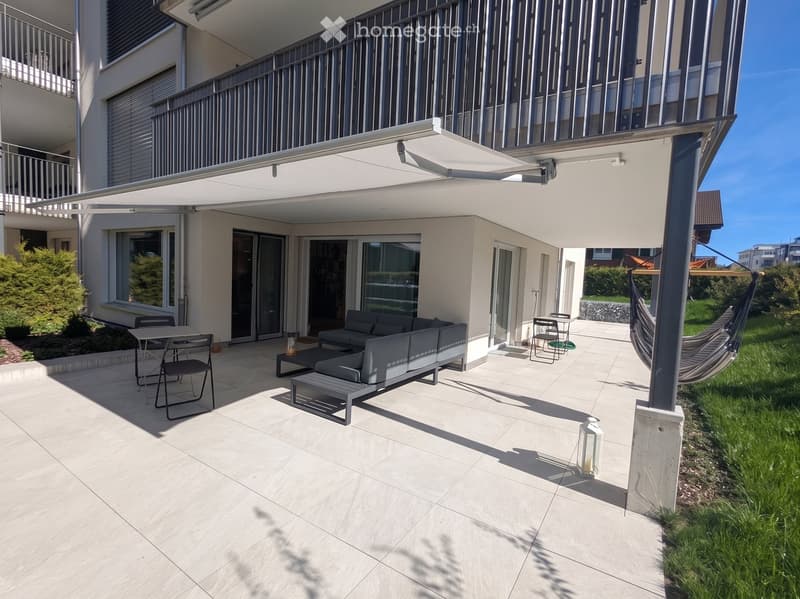 Modern outdoor patio with light-colored tiles, grey sectional sofa, small table, chairs, and a hammock.