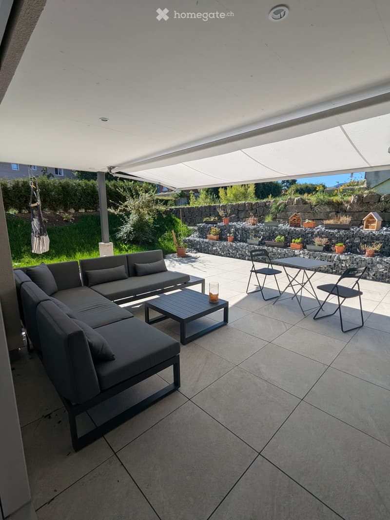 Outdoor patio with a large white canopy, grey sectional sofa, coffee table, folding chairs, and a small table with a glass candle holder. The patio is tiled and overlooks a landscaped garden with potted plants and a stone wall.