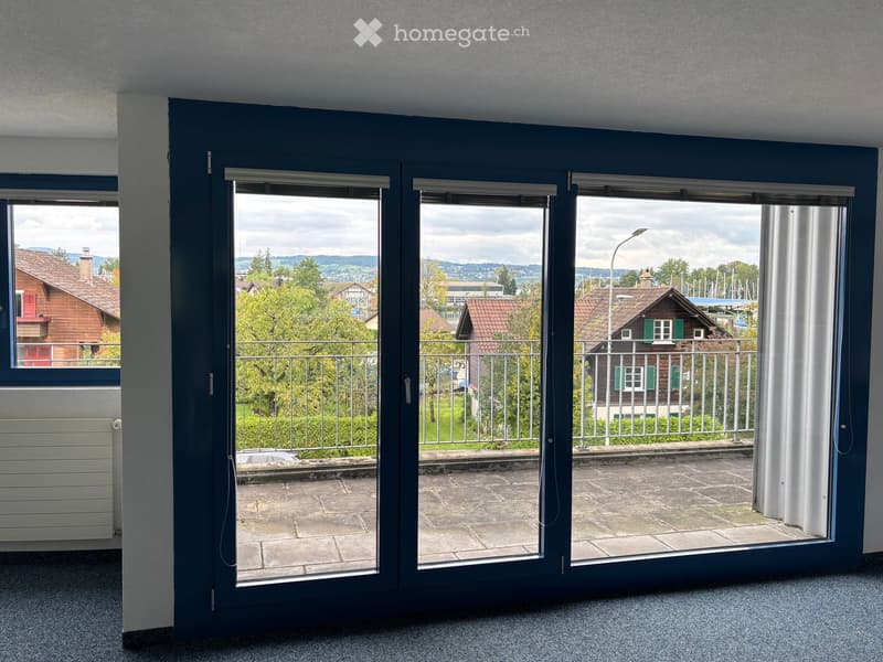 Balcony with large glass doors opening to a paved terrace, overlooking a scenic view of houses, trees, and a body of water.