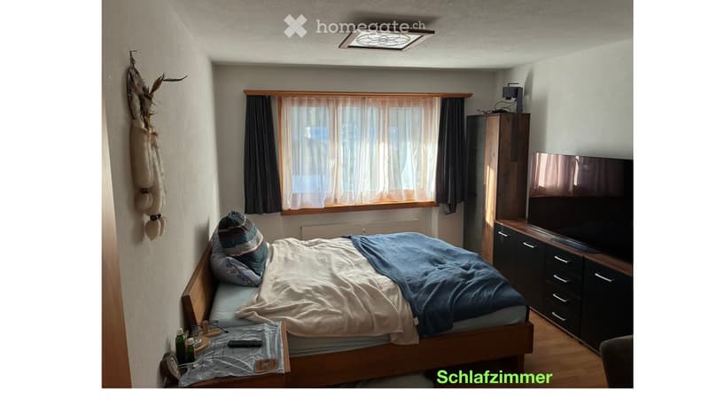 Cozy bedroom with a wooden bed, nightstand, television, and cabinet. The room features a window with curtains and a decorative wall hanging.