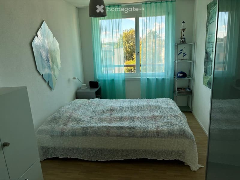 Cozy bedroom with a large bed, nightstand, wall-mounted lamp, decorative mirror, and a small shelf with various items.