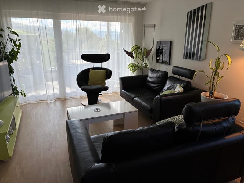 Modern living room with a black leather sofa, a black chair, a white coffee table, and several potted plants. The room features large windows with sheer curtains and a view of greenery outside.