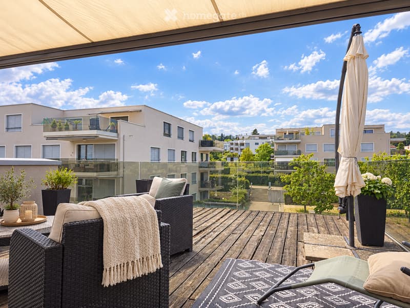 Balcony with wooden decking, wicker sofa, chair, small table, umbrella, and potted plants, overlooking a modern residential complex.