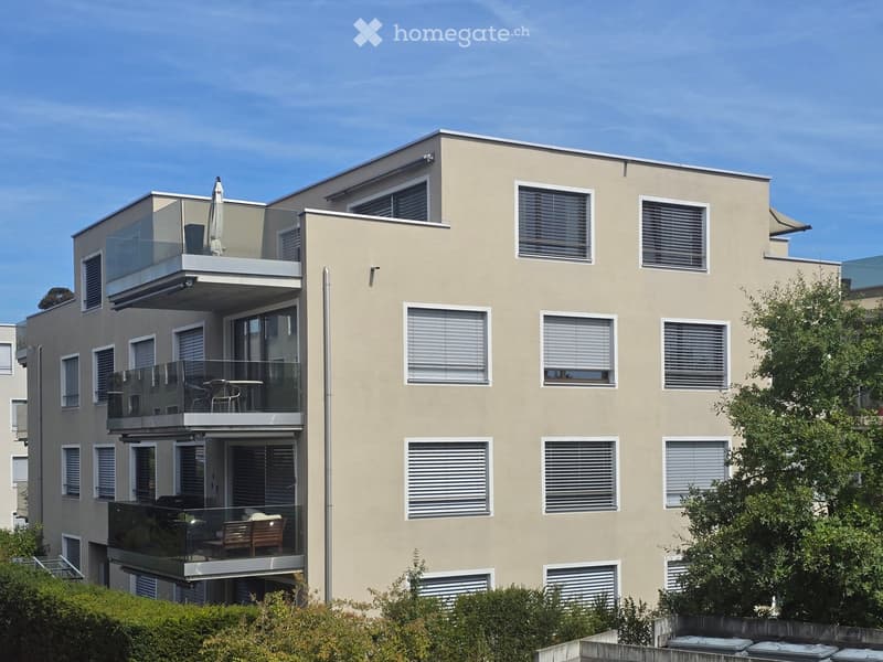 Modern multi-story building with beige facade, multiple balconies with glass railings, and outdoor furniture visible on some balconies.