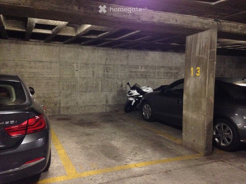 Underground parking area with concrete walls and ceiling, marked parking spaces, and several parked vehicles including a motorcycle and cars.