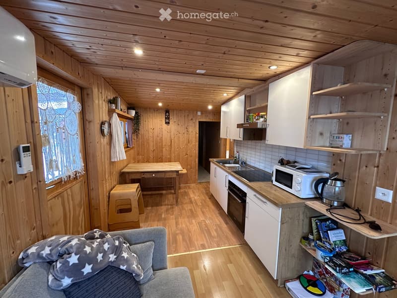 Cozy wooden interior with a sofa, small table, stool, microwave, oven, sink, fridge, and shelves with books and items.