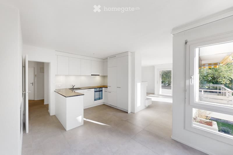Modern, minimalist kitchen with white cabinets, a central island, and a sink. The space is bright with large windows and a view of greenery outside.