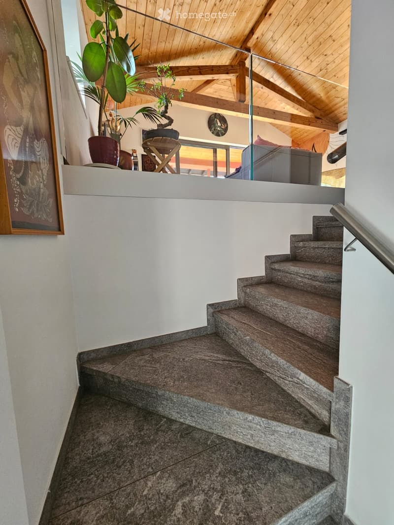 Staircase with stone steps and wooden handrail, leading to an upper level with a modern interior featuring exposed wooden beams and large windows.