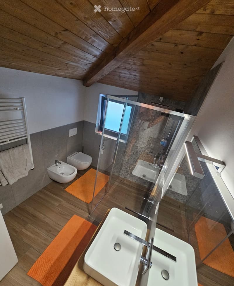 Modern bathroom with wooden flooring, a double sink, toilet, bidet, and a glass-enclosed shower area. Features include a towel rack with a towel and an orange bathroom mat.