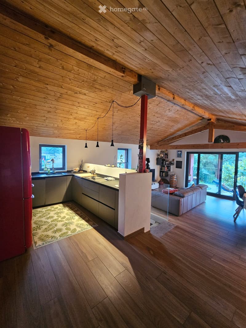 Open-plan living and kitchen area with wooden floors, exposed wooden beams, modern kitchen with stainless steel appliances, a red refrigerator, a grey sectional sofa, and a dining area with a table and chairs.