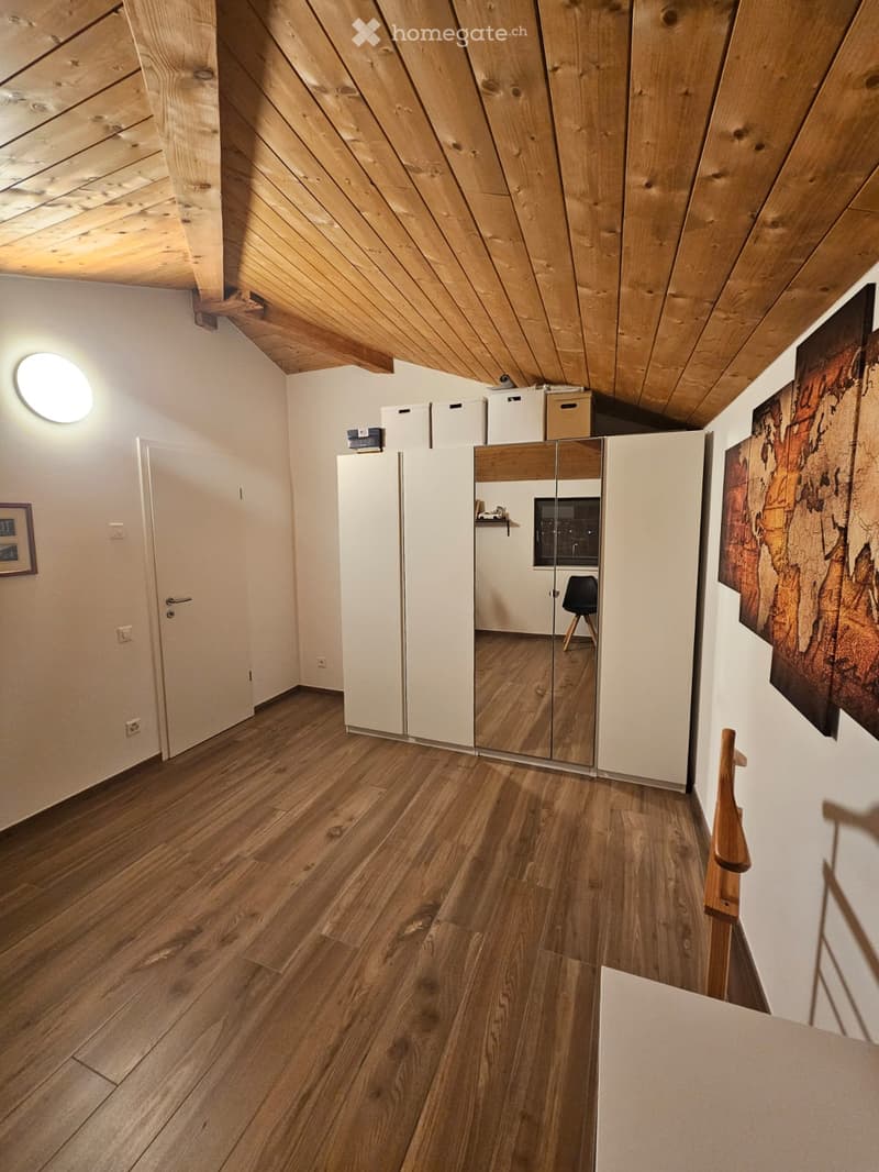 Bright hallway with wooden flooring, white walls, and a slanted wooden ceiling. Features a large mirrored wardrobe, a small black chair, and a round ceiling light.