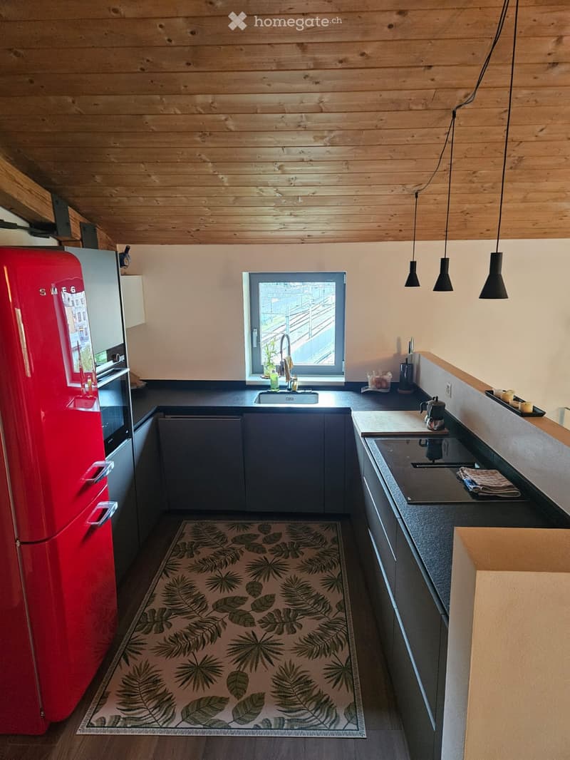 Modern kitchen with wooden ceiling, dark countertops, red refrigerator, sink, stove, and a leafy patterned rug.