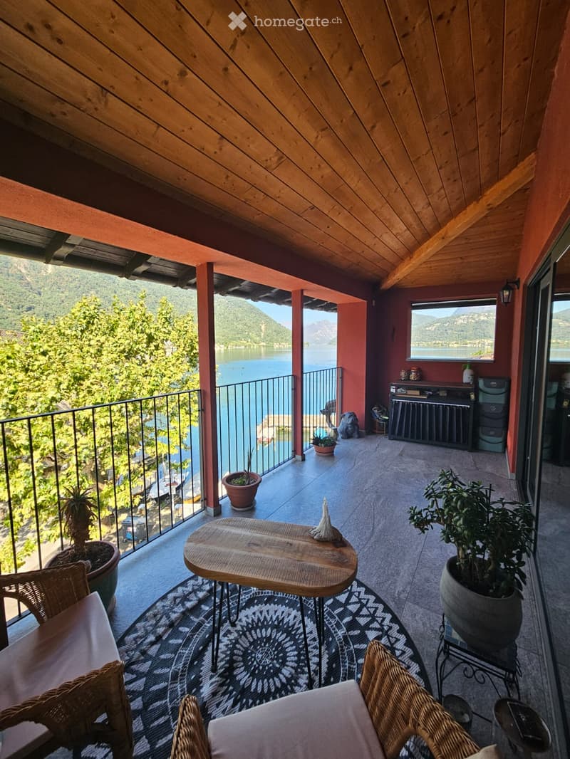 Balcony with wooden ceiling, tiled floor, and scenic lake view. Includes a small wooden table, wicker chairs, and potted plants.