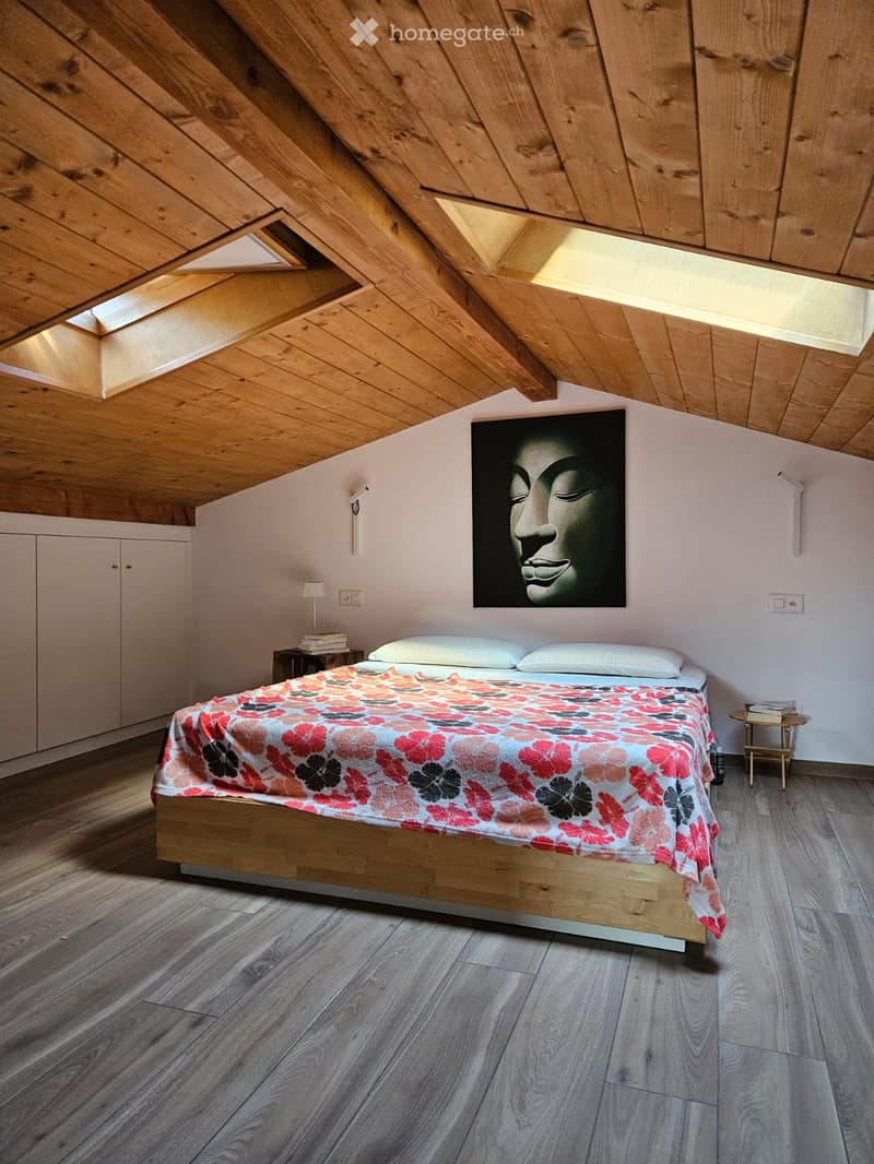 Modern attic bedroom with wooden slanted ceilings, skylights, a bed with a colorful floral duvet, nightstands, a lamp, and a large picture on the wall.
