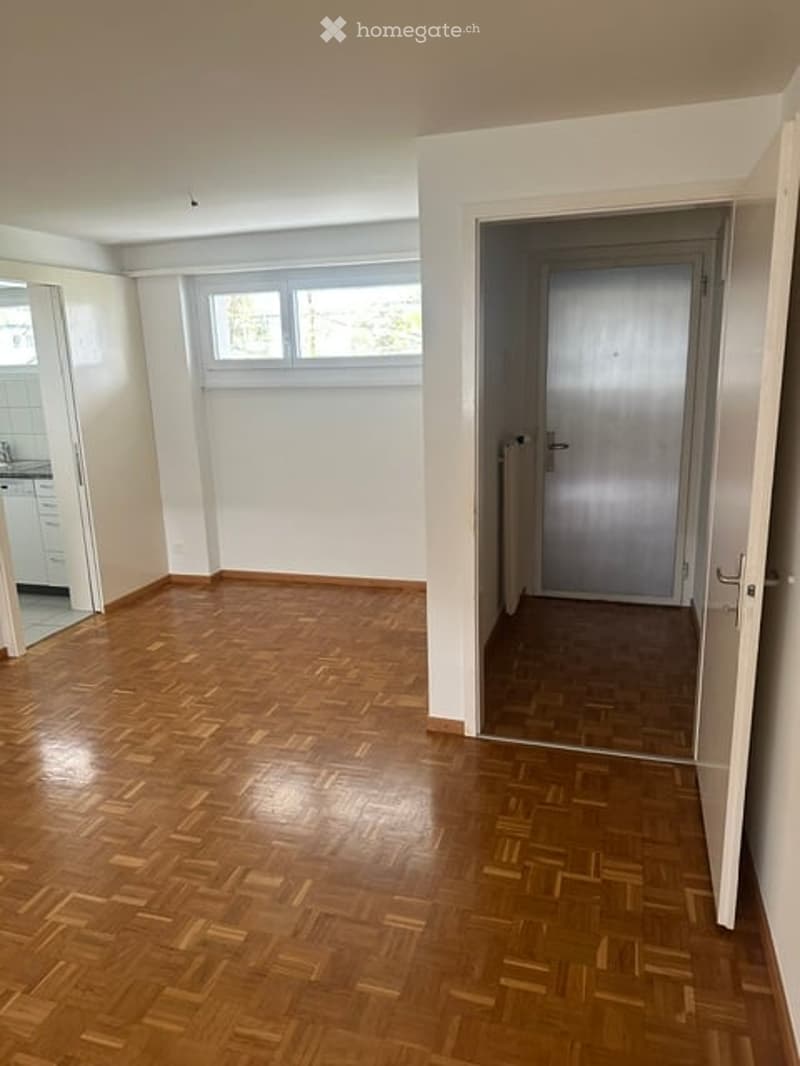 Empty room with wooden parquet flooring, white walls, and a large window letting in natural light. Adjacent room visible through an open doorway.