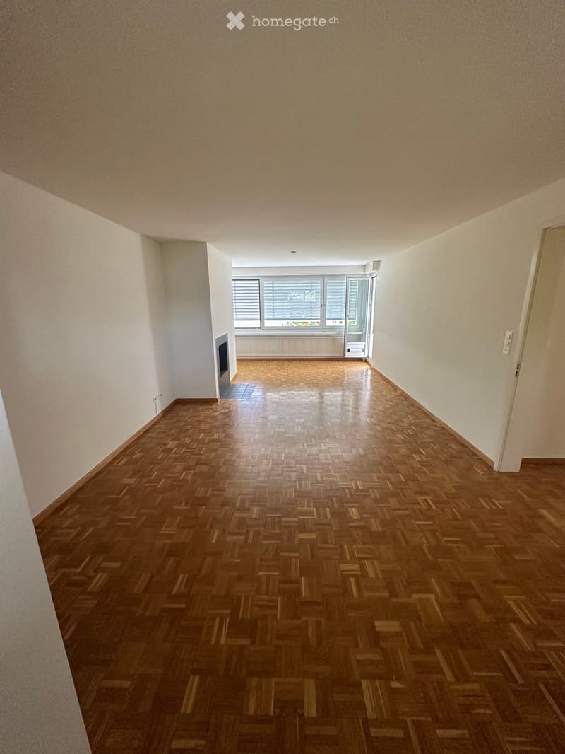 Empty hallway with wooden parquet flooring, white walls, and a large window with blinds at the end.